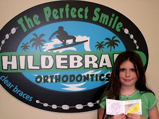 Woman smiling next to Hildebrand Orthodontics logo sign featuring tropical beach theme.