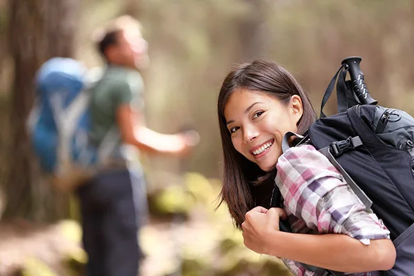 Young woman smiling at camera while hiking outdoors with backpack and friends in background.