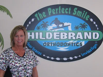 Woman smiling in front of Hildebrand Orthodontics "Perfect Smile" logo sign in office.