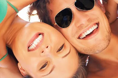 Two people smiling at camera wearing sunglasses in sunny outdoor setting, promoting dental health and orthodontics.