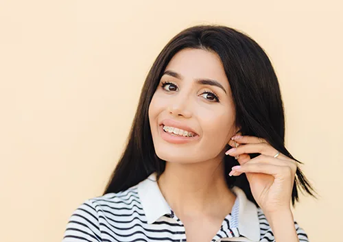 Headshot of cute lovely woman has broad smile, dark hair, keeps hand on ear, shows white teeth with braces, looks directly into camera, has happy expression, isolated over studio background.