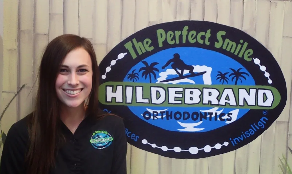 Young woman smiling next to Hildebrand Orthodontics blue oval logo sign indoors.