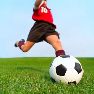 Young soccer player in red jersey kicking ball on grass field outdoors.