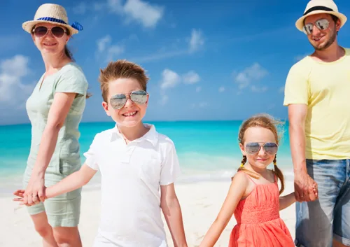 Family of four wearing sunglasses and hats smiling on sunny beach with turquoise ocean.
