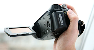 Hand holding compact video camera with flip-out screen in bright setting.