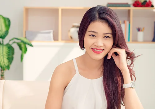 Smiling woman with long dark hair in modern home interior setting, touching her face.