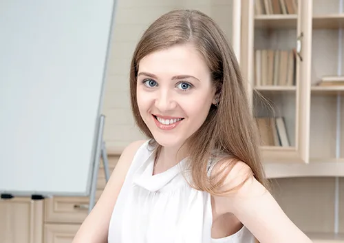 Young woman smiling at camera in bright office setting with whiteboard and shelving behind her.