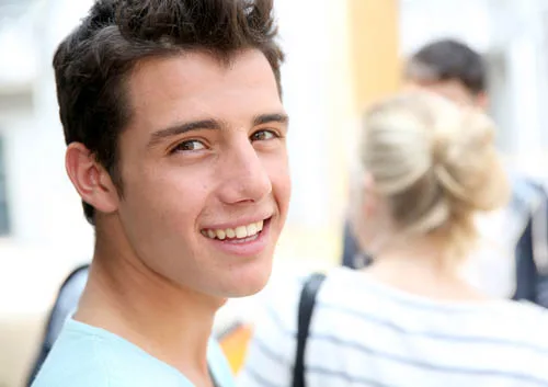 Young man smiling at camera in orthodontic office with blonde woman blurred in background.