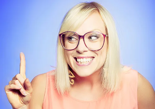 Young woman with blonde hair wearing glasses and pink top, smiling and pointing upward against blue background.