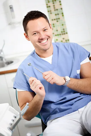 Smiling orthodontist in blue scrubs gesturing in modern dental office setting.