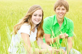 Two smiling young people sitting in green grass field outdoors during daytime.