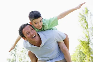 Father giving piggyback ride to smiling son outdoors in sunny setting.