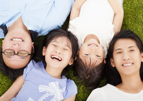 Four smiling people lying on green grass, looking down at camera in circle formation.