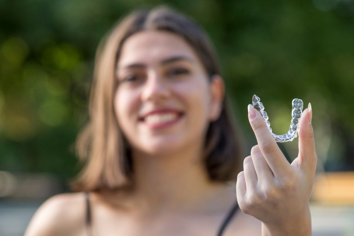 Young woman smiling outdoors, displaying teeth with hand gesture near face, orthodontic treatment showcase.