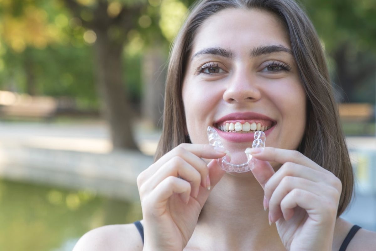 Young woman holding clear aligner outdoors, smiling at camera with blurred green park background.
