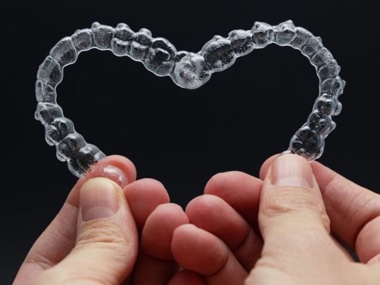 Two hands holding clear orthodontic aligner shaped like a heart against black background.