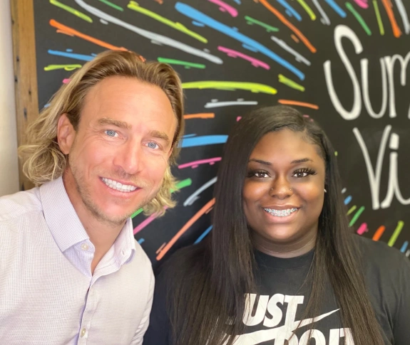 Two people smiling at camera in front of colorful orthodontics office backdrop.